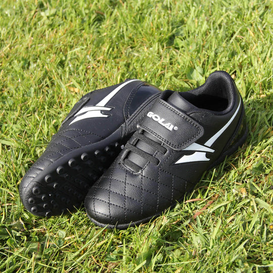 Astro turf boots. Black football trainers with stitching detail to the toes to give a quilted appearance. The touch close fastening and elasticated lace detail to the front. White Gola logo to the side and white Gola branding to the touch fasten strap. Black sole with small Astro turf studs to the base. Lifestyle photo of both feet on a field with right foot on its side to show sole.