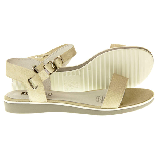 Beige sandals flat. Classic womens strappy sandals with beige faux leather straps around the ankle and over the toes. The ankle strap has a gold buckle fastening. Beige faux leather cushioned insoles. Very small wedge heel with beige outsole with a white rim around the top. Both feet from a side profile with left foot on its side behind the right to show the sole.