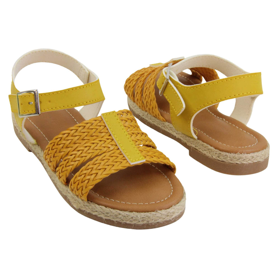 Big kids sandals. Triple strap sandals with a faux leather mustard yellow upper. Three woven faux leather straps over the foot with a plain band down the middle. A plain ankle strap with silver buckle. Brown insole with white lining. Brown outsole with jute rope style rim around the outside. Both feet at a slight angle facing top to tail.