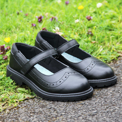 Black school shoes for teenage girls. Mary Jane style shoes with black faux leather uppers. With a touch fasten strap over the foot and brogue detailing around the top of the shoe. Black textile lining with a metallic blue insole. Chunky black sole with slight heel. Lifestyle photo with both shoes together at an angle half on some grass, half on a path.