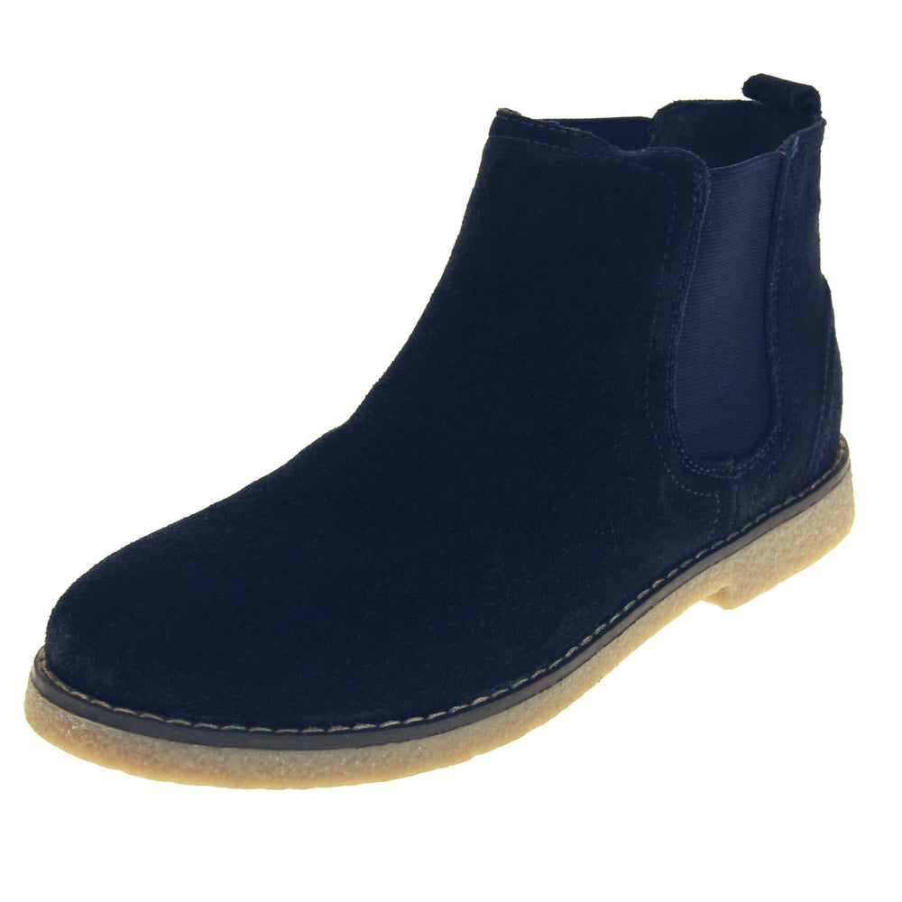 Blue suede ankle boots. Womens Chelsea boot style with a navy blue suede upper. Navy elasticated panels at the ankles and a navy loop at the heel to help pull them on. Beige coloured sole with a very slight heel. Left foot at an angle.