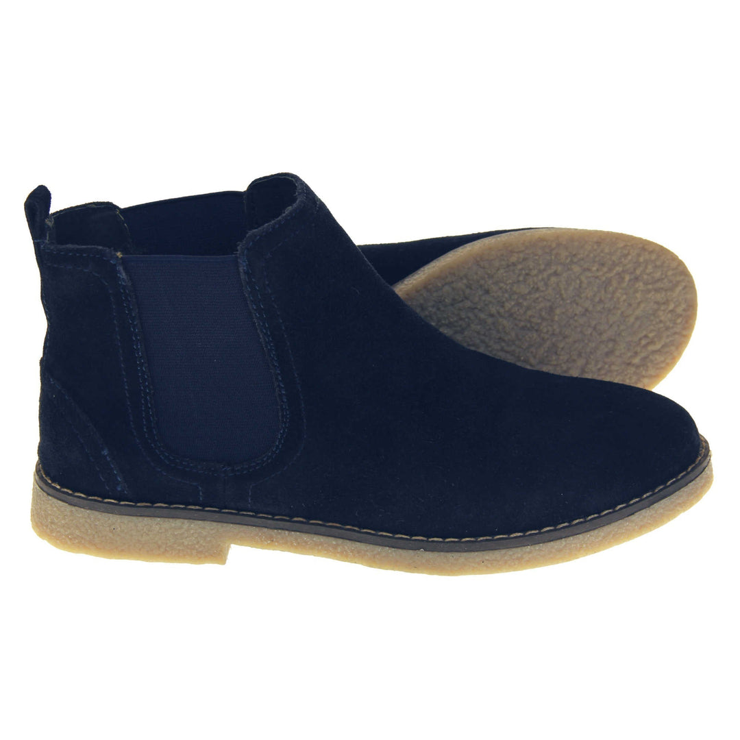 Blue suede ankle boots. Womens Chelsea boot style with a navy blue suede upper. Navy elasticated panels at the ankles and a navy loop at the heel to help pull them on. Beige coloured sole with a very slight heel. Both feet from a side profile with the left foot on its side to show the sole.