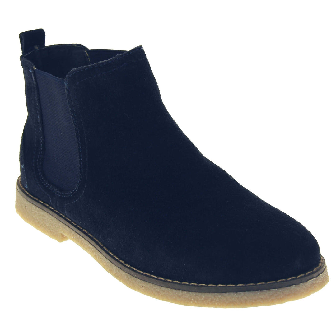 Blue suede ankle boots. Womens Chelsea boot style with a navy blue suede upper. Navy elasticated panels at the ankles and a navy loop at the heel to help pull them on. Beige coloured sole with a very slight heel. Right foot at an angle.