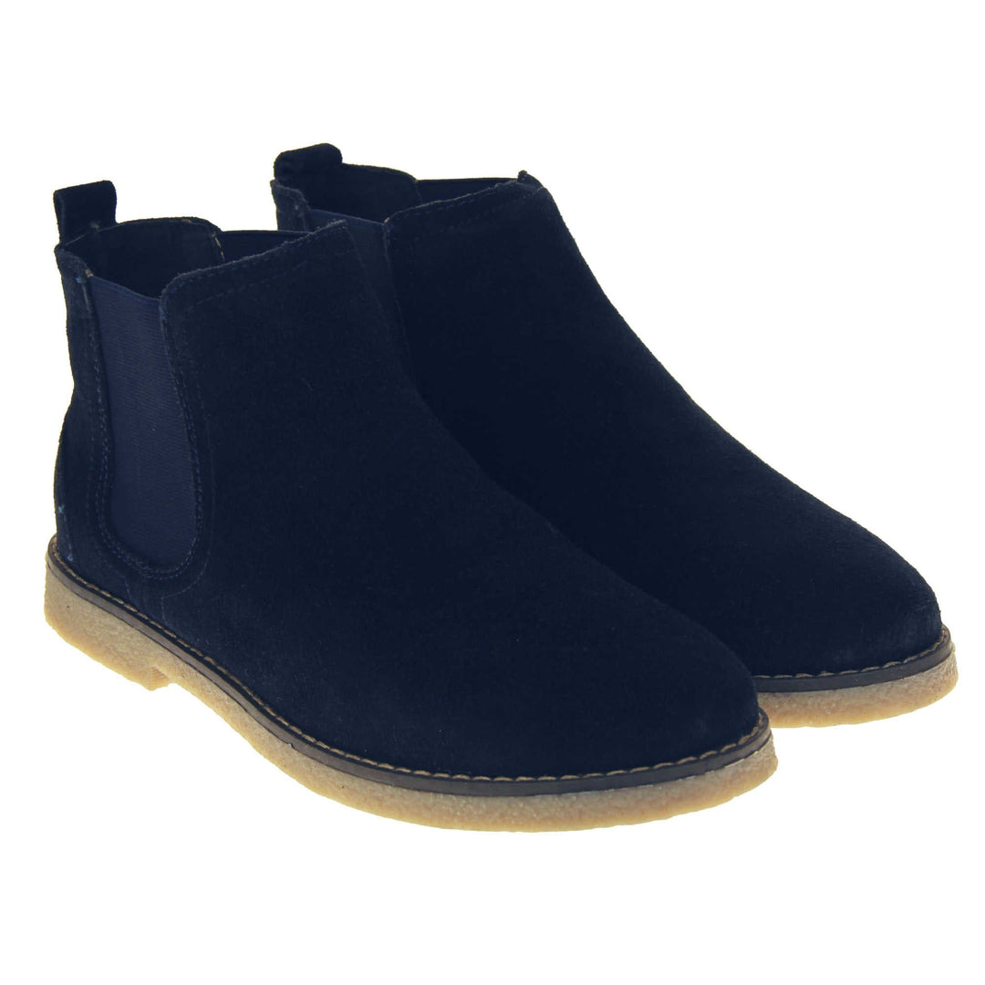 Blue suede ankle boots. Womens Chelsea boot style with a navy blue suede upper. Navy elasticated panels at the ankles and a navy loop at the heel to help pull them on. Beige coloured sole with a very slight heel. Both feet together from an angle.