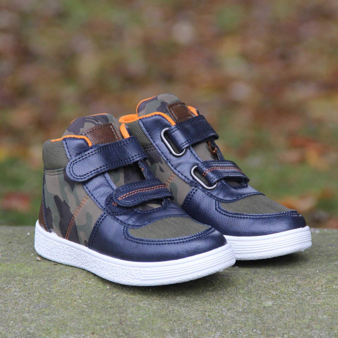 Boots for Boys. Boys canvas ankle boots with camouflage sides and tongue, khaki collar and upper toes and navy faux leather edging details. Touch fasten straps to the front with a white sole. Lifestyle shot with both feet on a wall in the park