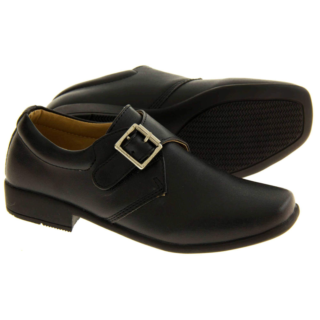 Boys Formal Shoes. Black matt monk style shoes with a buckle effect touch close fastening and a black sole. Both feet with the left foot on its side to show the sole