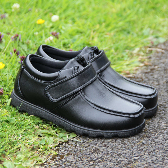 Boys smart school shoes. Black low top shoes with faux leather uppers. Stitching detail with a squared toe and a touch fasten strap over the top of the shoe. Black textile lining with light blue insole and black sole with chunky grip. Lifestyle photo with both shoes together at an angle half on some grass, half on a path.
