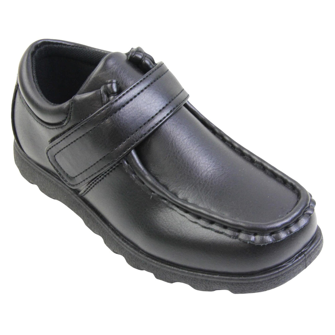 Boys smart school shoes. Black low top shoes with faux leather uppers. Stitching detail with a squared toe and a touch fasten strap over the top of the shoe. Black textile lining with light blue insole and black sole with chunky grip. Right foot at an angle.