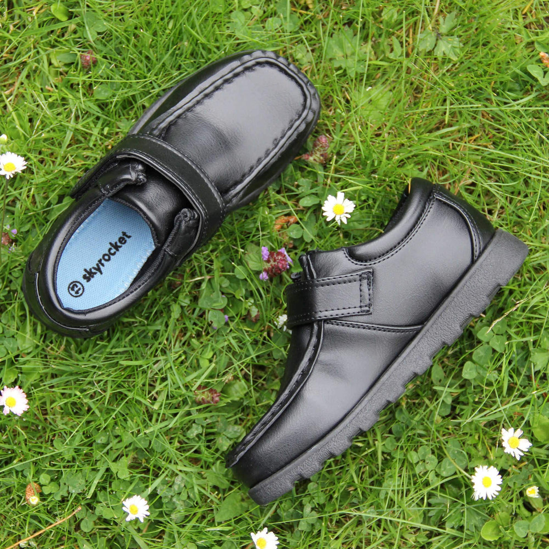 Boys smart school shoes. Black low top shoes with faux leather uppers. Stitching detail with a squared toe and a touch fasten strap over the top of the shoe. Black textile lining with light blue insole and black sole with chunky grip. Lifestyle photo with both shoes on grass from above facing top to tail with one on its side.