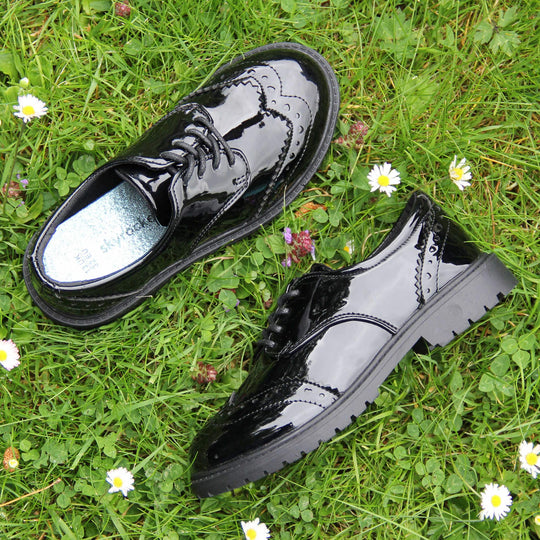 Brogue school shoes. Black patent faux leather uppers in a brogue style shoe. Black laces and lining with metallic blue insole. Black sole with very slight heel. Lifestyle photo with both shoes on some grass from above facing top to tail with one on its side.