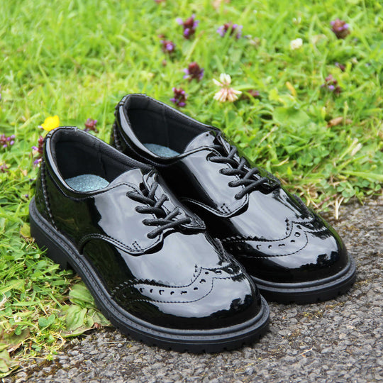 Brogue school shoes. Black patent faux leather uppers in a brogue style shoe. Black laces and lining with metallic blue insole. Black sole with very slight heel. Lifestyle photo with both shoes together at an angle on some grass.