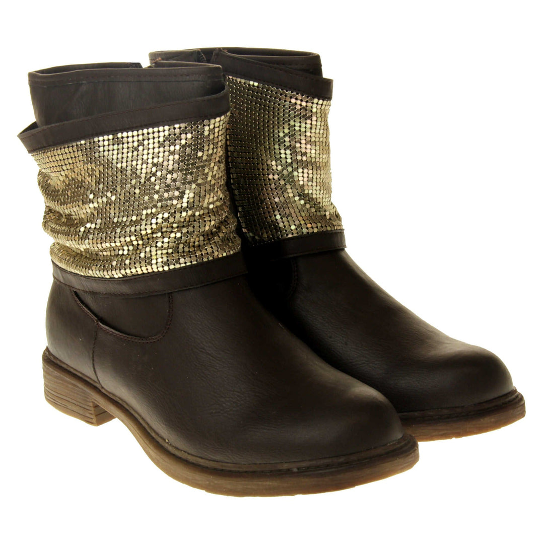 Brown faux leather ankle boots. Biker style boots with a dark brown upper. A thick band of shiny gold chainmail runs around the ankle. Zip fastening down the inside of the boot. Brown sole with a slight heel. Both feet together from an angle.