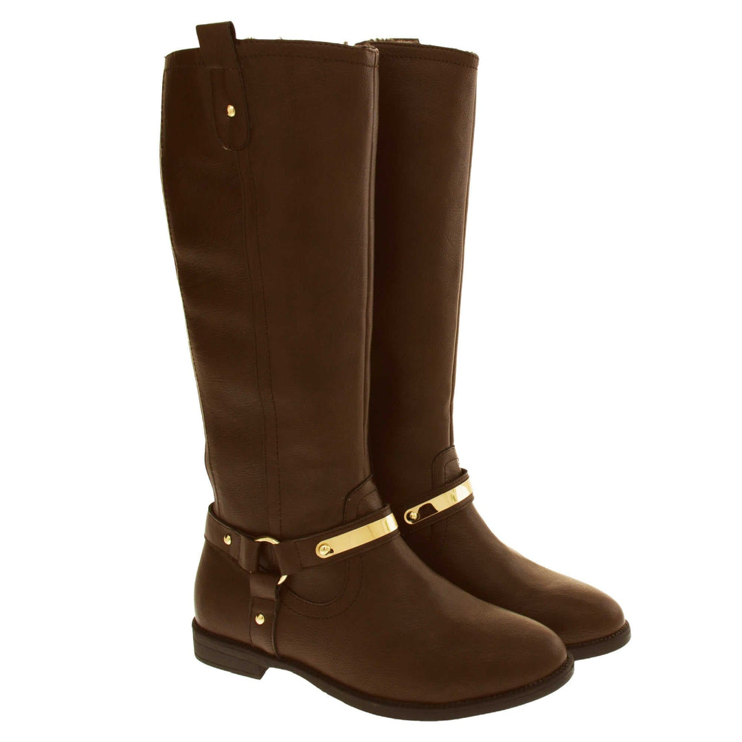 Brown leather boots women. Knee high style boots with a dark brown faux leather upper. A Tri strap going around the ankle and down to the bottom of the boot joined by the ankle with a good loop. Gold plate to the front of the ankle strap. Zip up the inside of the boot. Faux leather loop on the top of the outside of the boot to help with pulling it on. Both shoes together at an angle.