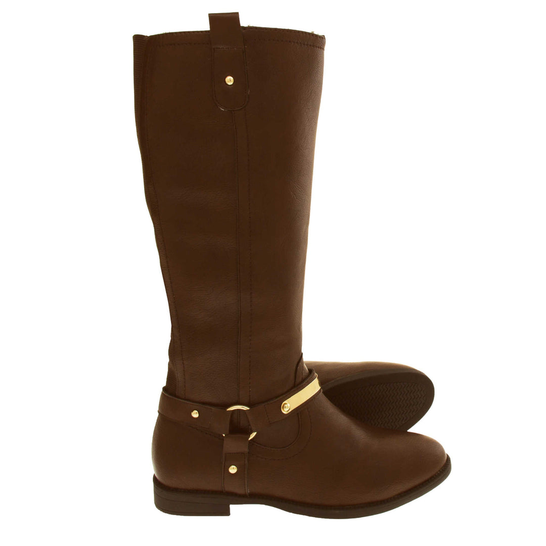 Brown leather boots women. Knee high style boots with a dark brown faux leather upper. A Tri strap going around the ankle and down to the bottom of the boot joined by the ankle with a good loop. Gold plate to the front of the ankle strap. Zip up the inside of the boot. Faux leather loop on the top of the outside of the boot to help with pulling it on. Both feet from a side profile with the left foot on its side to show the sole.