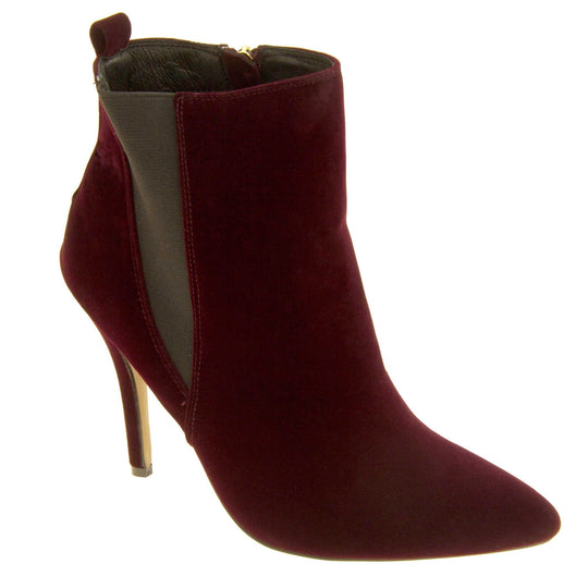 Burgundy stiletto ankle boots. Womens ankle boots with a dark red velvet upper. Grey elasticated gusset and a burgundy loop at the heel to help pull them on. Brown high stiletto heel and outsole with cream coloured bottom. Black textile lining and zip fastening to the inside of the leg. Right foot to the side.