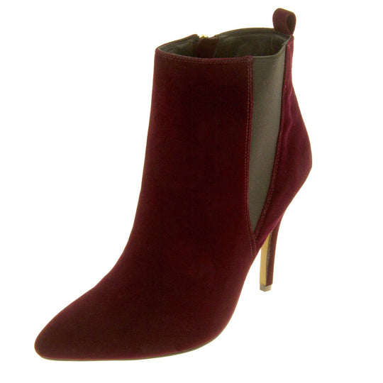 Burgundy stiletto ankle boots. Womens ankle boots with a dark red velvet upper. Grey elasticated gusset and a burgundy loop at the heel to help pull them on. Brown high stiletto heel and outsole with cream coloured bottom. Black textile lining and zip fastening to the inside of the leg. Left foot to the side.