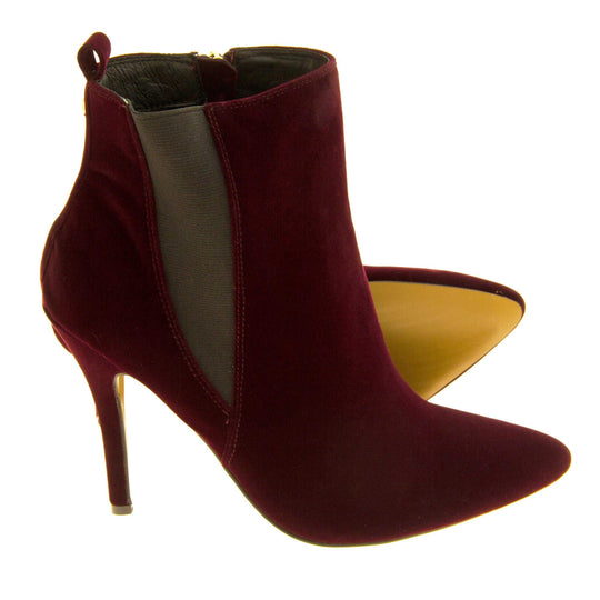 Burgundy stiletto ankle boots. Womens ankle boots with a dark red velvet upper. Grey elasticated gusset and a burgundy loop at the heel to help pull them on. Brown high stiletto heel and outsole with cream coloured bottom. Black textile lining and zip fastening to the inside of the leg. Both feet from a side profile with the left foot on its side to show the sole.