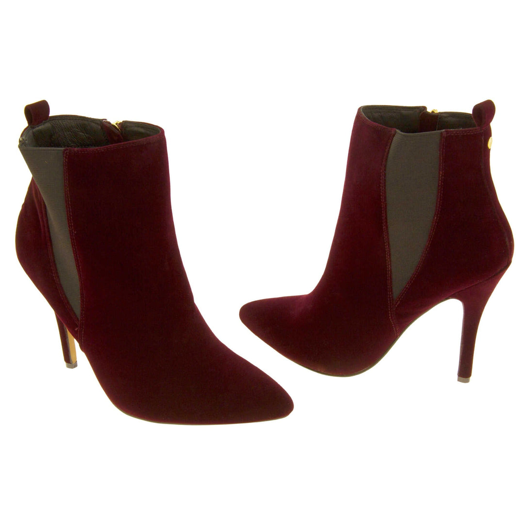 Burgundy stiletto ankle boots. Womens ankle boots with a dark red velvet upper. Grey elasticated gusset and a burgundy loop at the heel to help pull them on. Brown high stiletto heel and outsole with cream coloured bottom. Black textile lining and zip fastening to the inside of the leg. Both feet from a slight angle facing top to tail.