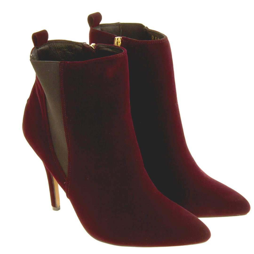 Burgundy stiletto ankle boots. Womens ankle boots with a dark red velvet upper. Grey elasticated gusset and a burgundy loop at the heel to help pull them on. Brown high stiletto heel and outsole with cream coloured bottom. Black textile lining and zip fastening to the inside of the leg. Both feet together from an angle.