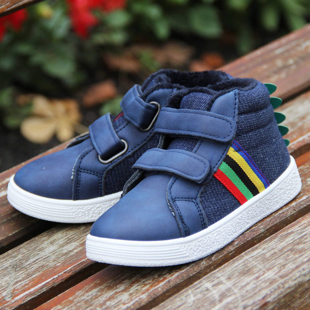 Kids Denim Blue Hi-top Trainer Boots - Denim blue and smooth faux leather upper with dual hook and loop fastening, white outsole, rainbow stripe to side, green dinosaur spine to heel and dark blue fleece lining. Both feet taken in autumn shot on bench.