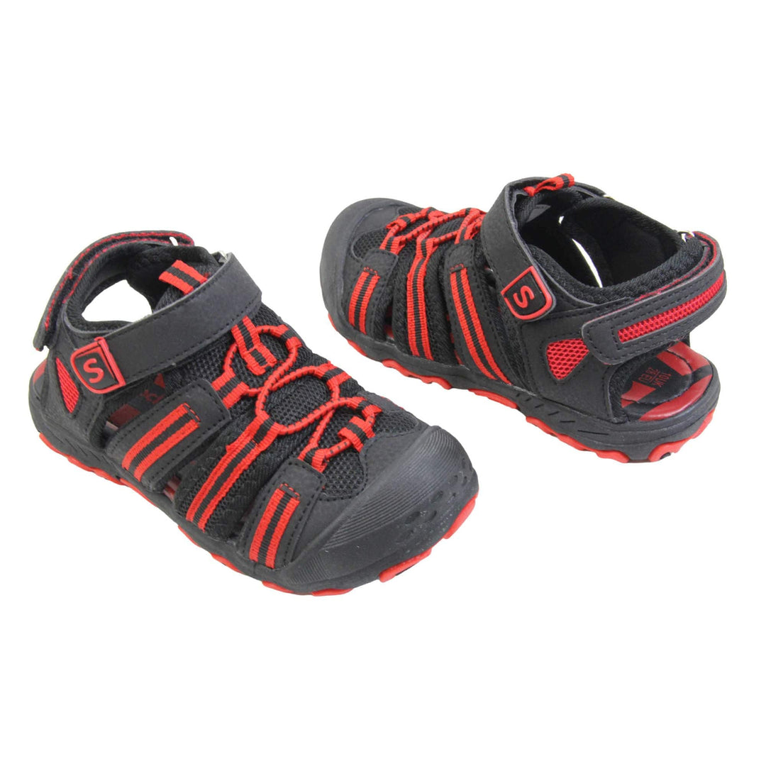 Closed toe kids sandals. Strappy beach sandals with black mesh and faux leather uppers. With black and red textile strap detailing. Red elasticated faux laces to the front. Black faux leather straps around the front and back of the ankle with touch fastening. Red insole and black rubbery outsole and toe bumper with black and red grip on the bottom. Both feet at an angle facing top to tail.