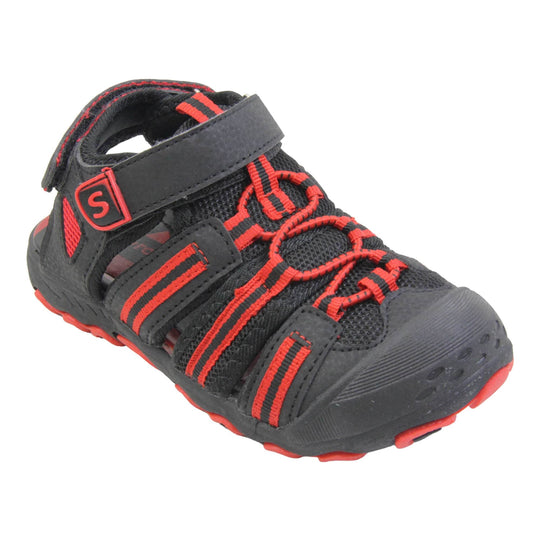 Closed toe kids sandals. Strappy beach sandals with black mesh and faux leather uppers. With black and red textile strap detailing. Red elasticated faux laces to the front. Black faux leather straps around the front and back of the ankle with touch fastening. Red insole and black rubbery outsole and toe bumper with black and red grip on the bottom. Right foot at an angle.