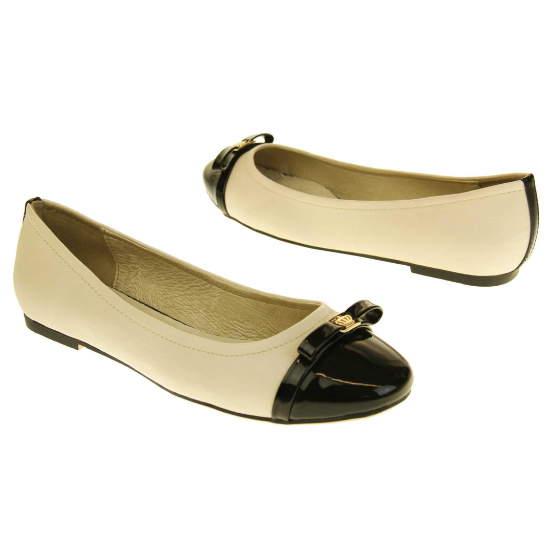 Cream dolly shoes. Cream leather ballet pumps with black patent toe and bow detail. Black stripe down the heel of the upper. Beige outsole with black rim and with a very slight heel. Both feet at an angle facing top to tail.