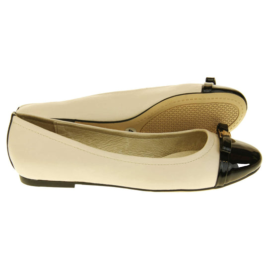 Cream dolly shoes. Cream leather ballet pumps with black patent toe and bow detail. Black stripe down the heel of the upper.Beige outsole with black rim and with a very slight heel.  Both feet from a side profile with the left foot on its side behind the the right foot to show the sole.