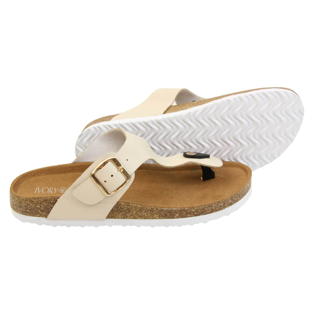 Cream flip flops. Cream faux leather strap with toe post to the front and gold buckle to the outside. Soft tan faux suede footbed with cork effect outsole and white sole. Both feet from a side profile with the left foot on its side behind the the right foot to show the sole.