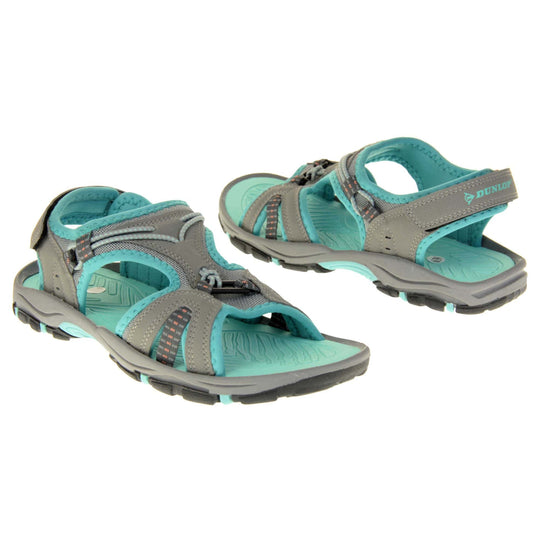 Dunlop womens walking sandals - Light grey faux leather upper with decorative elasticated strap detailing and teal blue trim. Hook and loop touch fastening ankle strap for secure fitting. Turquoise padded insole for extra comfort and a teal, grey and black flexible outsole with good grips for hiking. Both feet facing different directions.