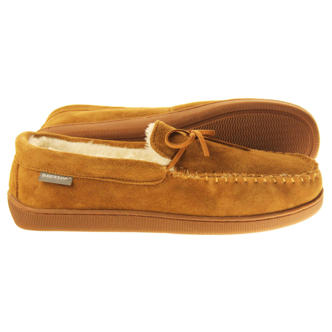 Faux fur moccasins. Closed back slippers in a moccasin style with tan brown suede leather upper and bow. Cream faux fur lining. Thick brown sole. Both feet from a side profile with the left foot on its side to show the sole.