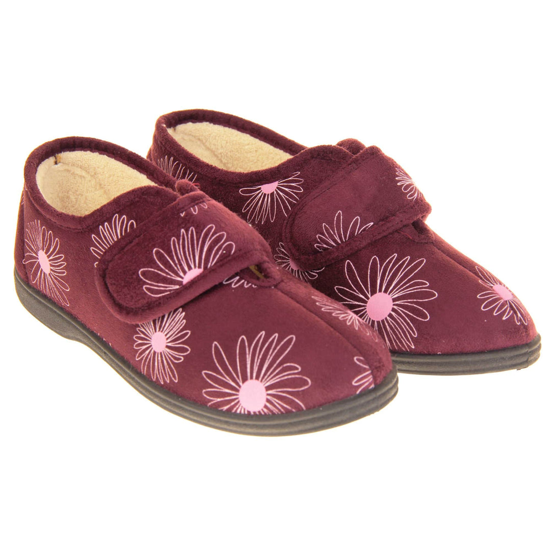 Floral slippers. Womens full back slipper. Burgundy upper with white daisy design and bright pink for the middle of the flowers. Touch fasten strap over the top of the foot to adjust the fit. Cream fleece lining and firm black sole. Both feet together at an angle.