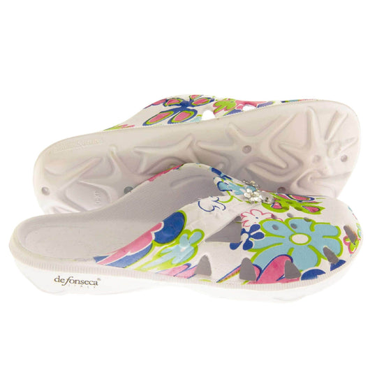 Girls Clogs. White synthetic clogs in a mule style. Cut out holes around the toes and the upper. Bright, fun floral print on the upper with diamante detailing to the centre. Matching white sole. Both feet from side profile with left foot on its side to show the sole.