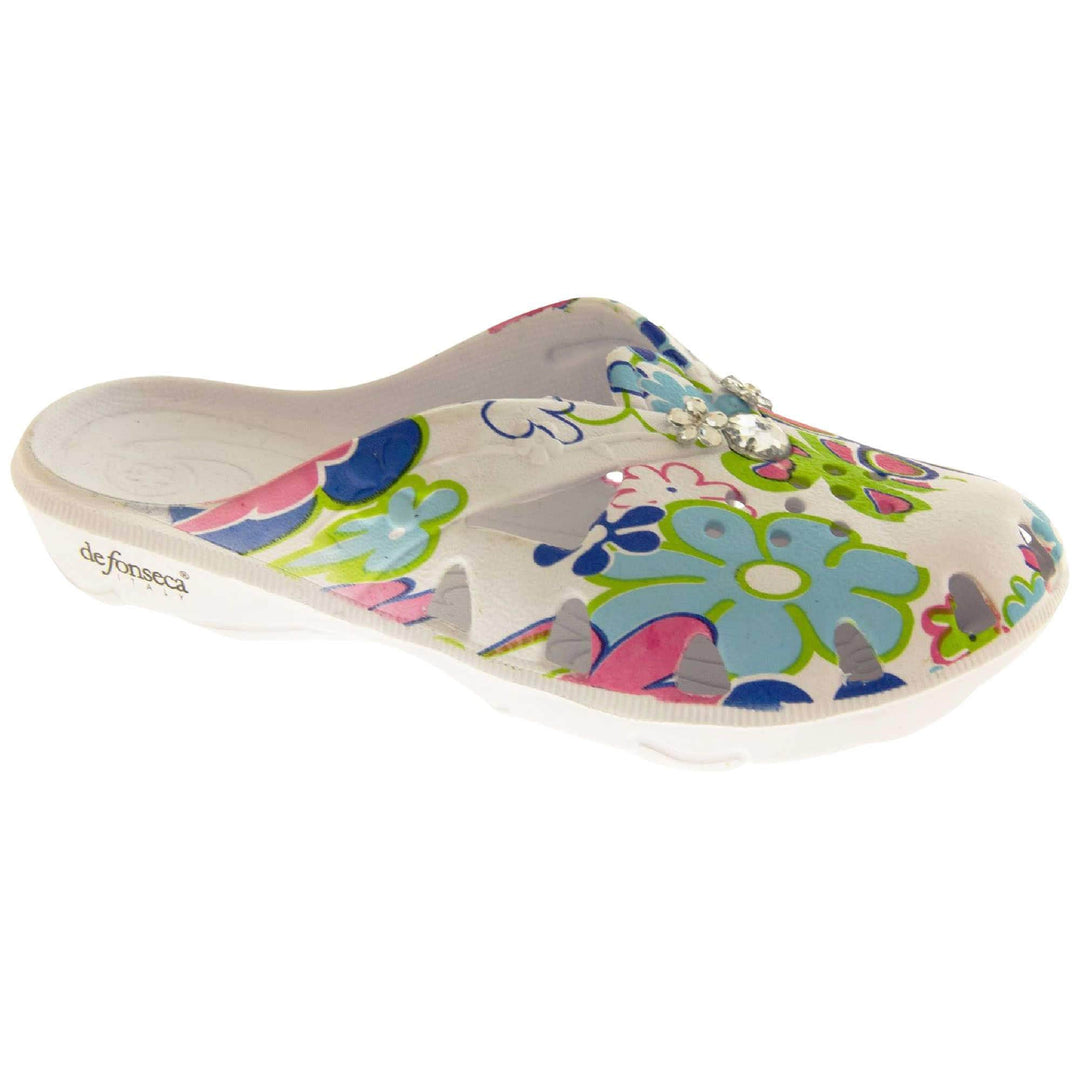 Girls Clogs. White synthetic clogs in a mule style. Cut out holes around the toes and the upper. Bright, fun floral print on the upper with diamante detailing to the centre. Matching white sole. Right foot at an angle.