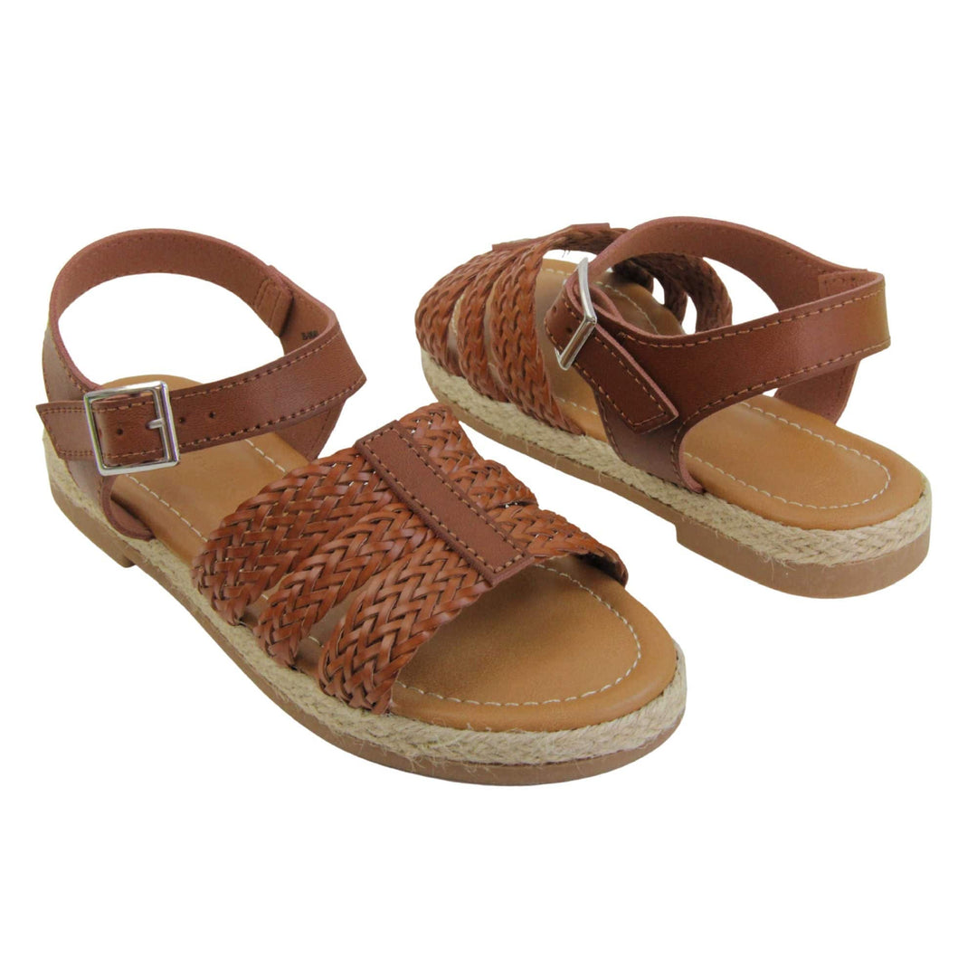 Girls flat sandals. Triple strap sandals with a faux leather dark brown upper. Three woven faux leather straps over the foot with a plain band down the middle. A plain ankle strap with silver buckle. Brown insole with brown lining. Brown outsole with jute rope style rim around the outside. Both feet at an angle facing top to tail.