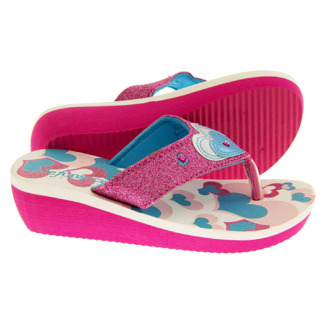 Girls glitter sandals. Foam wedge sandals for girls. Pink bottom half of the sole with ridges for grip, white top half with heart designs in pale pink, bright pink and teal. Bright pink glitter strap with toe-post. With cream and teal heart detail to the strap and pink diamantes. Both feet from a sidfe profile with the left foot on its side to show the sole.