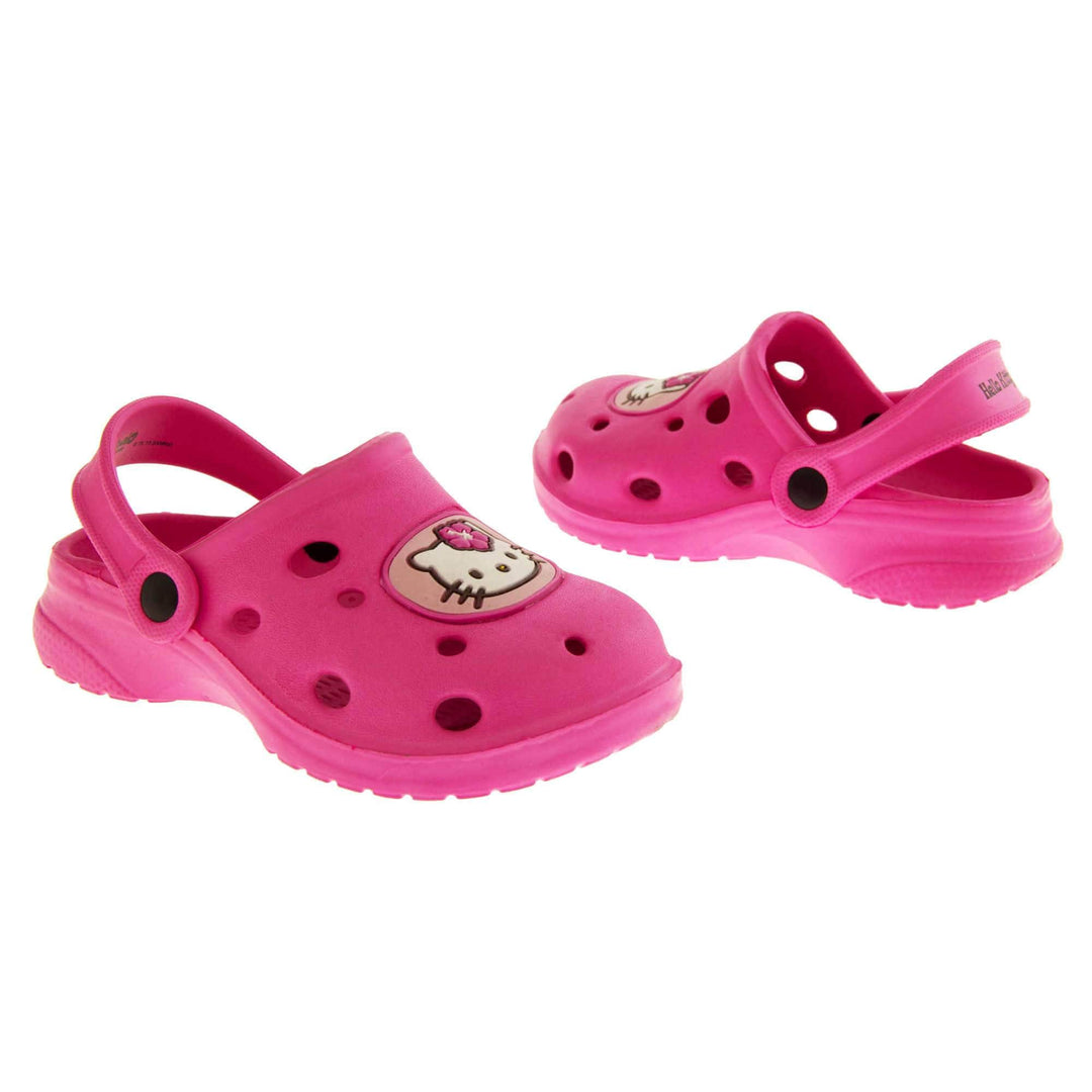 Girls Sandals Pink. Fuchsia pink synthetic clog sandals with holes in the upper. Hello Kitty face in a pale pink circle to the front. Pink strap with black studs to attach to the back of the shoe. The strap can be worn around the back of the heel or over the top of the shoe in a mule. Pink sole with slight heel to the back. Both shoes facing top to tail and from an angle.