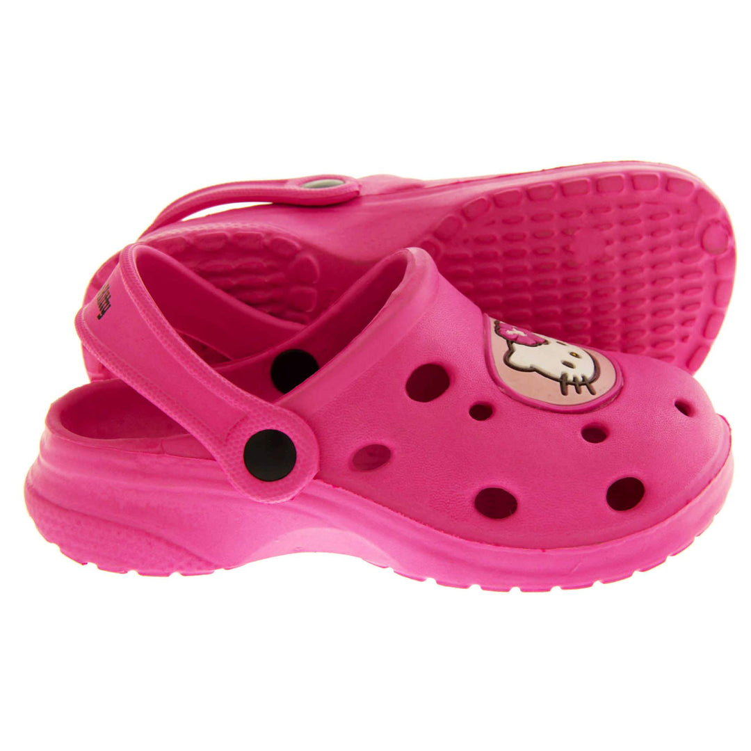 Girls Sandals Pink. Fuchsia pink synthetic clog sandals with holes in the upper. Hello Kitty face in a pale pink circle to the front. Pink strap with black studs to attach to the back of the shoe. The strap can be worn around the back of the heel or over the top of the shoe in a mule. Pink sole with slight heel to the back. Both feet from a side profile with the left foot on its side to show the sole.