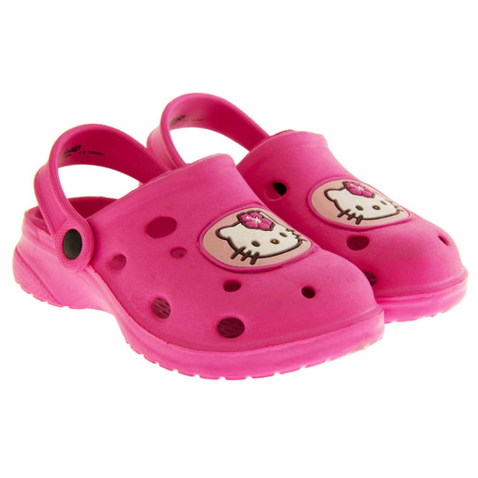 Girls Sandals Pink. Fuchsia pink synthetic clog sandals with holes in the upper. Hello Kitty face in a pale pink circle to the front. Pink strap with black studs to attach to the back of the shoe. The strap can be worn around the back of the heel or over the top of the shoe in a mule. Pink sole with slight heel to the back. Both feet next to each other at an angle.