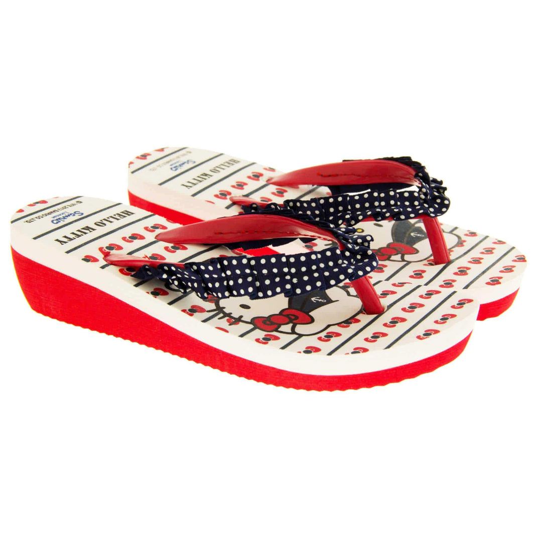 Girls wedge sandals. Hello Kitty flip flop sandal. White top with Hello Kitty design. Red synthetic between toe strap with blue textile frill with white spots. Small wedge heel. Red sole. Both feet next to each other at an angle.