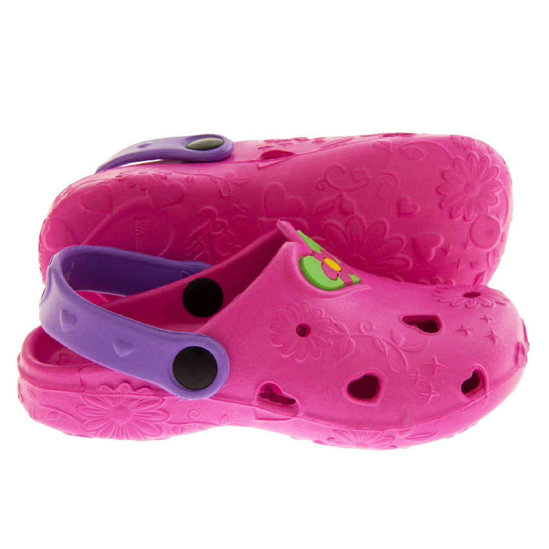 Girls Sandals. Pink synthetic clog style shoes with embossed flowers and stars. Cut out love hearts on the upper. Green love heart, with pink and yellow flower in the centre, to the middle top of the upper. Purple strap that goes along the back of your heel. The strap can be moved along the top of the shoe instead to make the shoe a mule. Both feet from side profile with left foot on its side to show the sole.