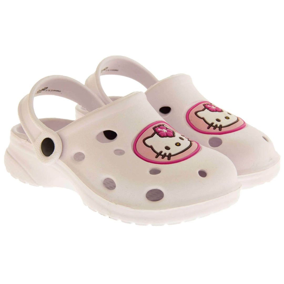Girls Sandals White. White synthetic clog sandals with holes in the upper. Hello Kitty face in a pink circle to the front. White strap with black studs to attach to the back of the shoe. The strap can be worn around the back of the heel or over the top of the shoe in a mule. White sole with slight heel to the back. Both feet next to each other at an angle.