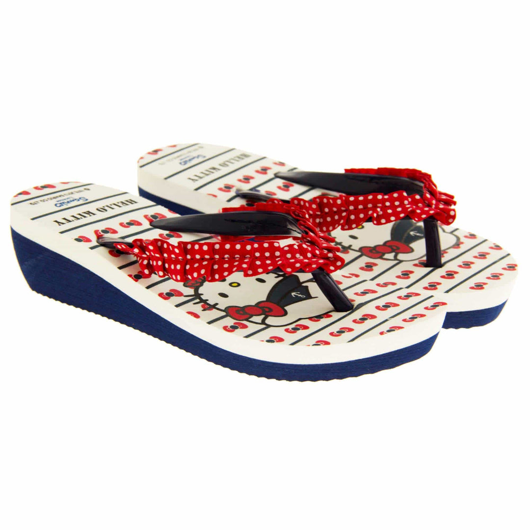 Girls wedge sandals. Hello Kitty flip flop sandal. White top with Hello Kitty design. Blue synthetic between toe strap with red textile frill with white spots. Small wedge heel. Blue sole. Both feet next to each other at an angle.