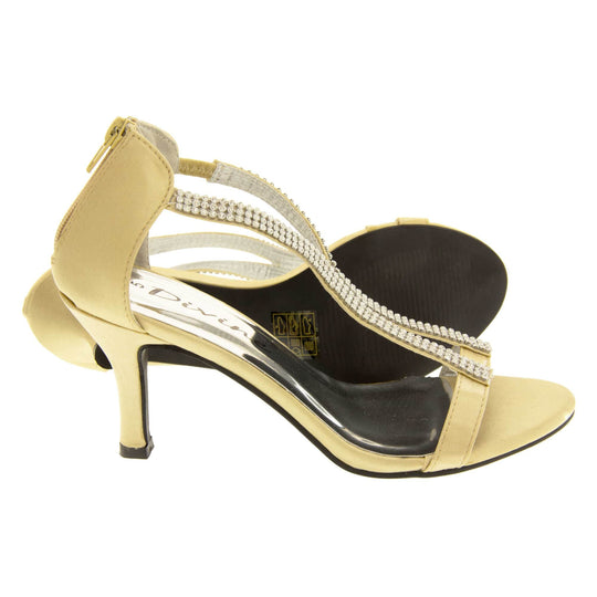 Gold strappy sandals. Womens high heels with open toes, two wavy gold satin straps covered in diamantes going down the front of the foot meeting at a plain gold satin toe strap. Closed back in the same gold satin with zip up the back for putting them on. Metallic silver insoles. Gold satin stiletto heel and black sole. Both feet from a side profile with the left foot on its side behind the the right foot to show the sole.