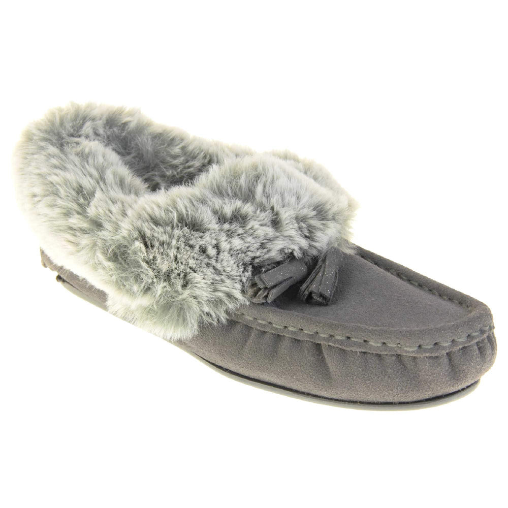 Grey Moccasin Slippers. Womens grey faux suede moccasin style slippers. With stitched detailing and tassels with subtle glitter on. Pale grey faux fur collar and lining. Grey firm sole. Right foot at an angle.