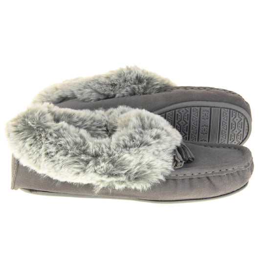 Grey Moccasin Slippers. Womens grey faux suede moccasin style slippers. With stitched detailing and tassels with subtle glitter on. Pale grey faux fur collar and lining. Grey firm sole. Both feet from a side profile with the left foot on its side behind the the right foot to show the sole.