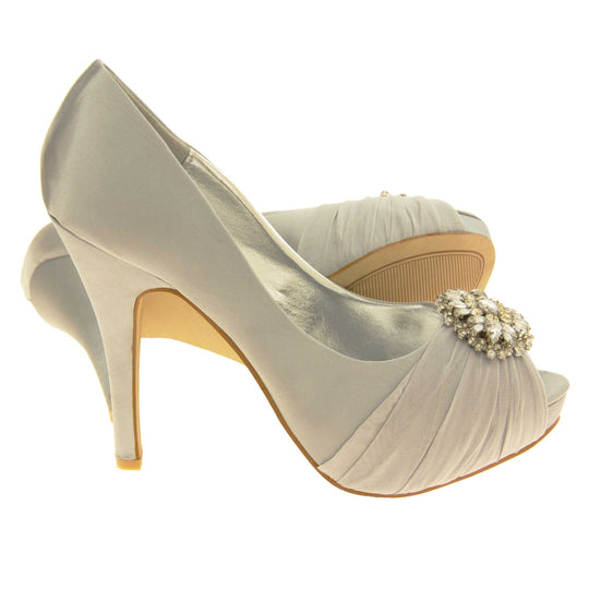 Grey satin heels. Classic women's peep toe high heels with a pale grey satin upper. Metallic silver insole with Sabatine branding. Grey satin stiletto heel with a cream sole. Diamante cluster and ruched detailing across the toes. Both feet from a side profile with the left foot on its side behind the the right foot to show the sole.