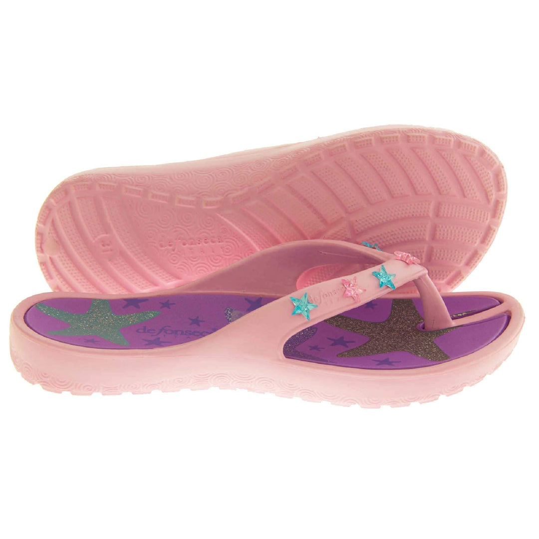 Kids flip flops girls. Pale pink flip flops with toe post strap. Purple insole with glittery heart design on. Small pink and teal plastic stars along the strap. Slip-resistant grip to the sole. Both feet from side profile with left foot on its side to show the sole.