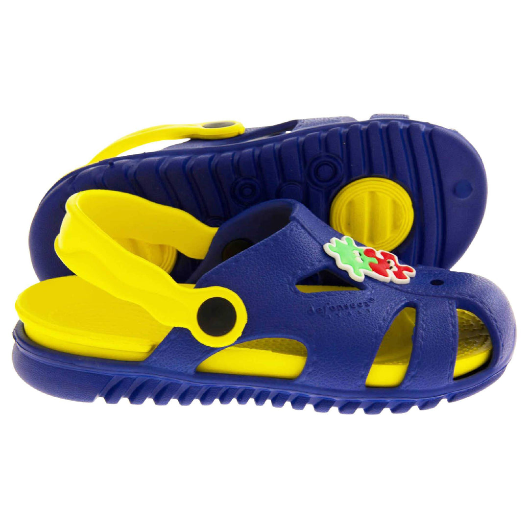 Kids pool clogs. Fisherman style synthetic sandals with blue upper and outer sole. Yellow ankle strap and insole. Green and red jigsaw pieces detail to the centre of the upper. Both feet from side profile with left foot on its side to show the sole.
