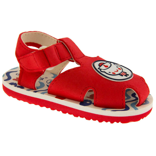 Boys Sandals. Fisherman style sandals with red upper and touch fasten ankle strap. The red upper has a white circle in the centre with a bear dressed as a sailor inside it. The inside of the upper and straps is white, as is the top half of the sole with the foot bed, which has a blue wave pattern on it. The bottom half of the sole is red and has a bumpy grip to the bottom of it. Right foot at an angle.